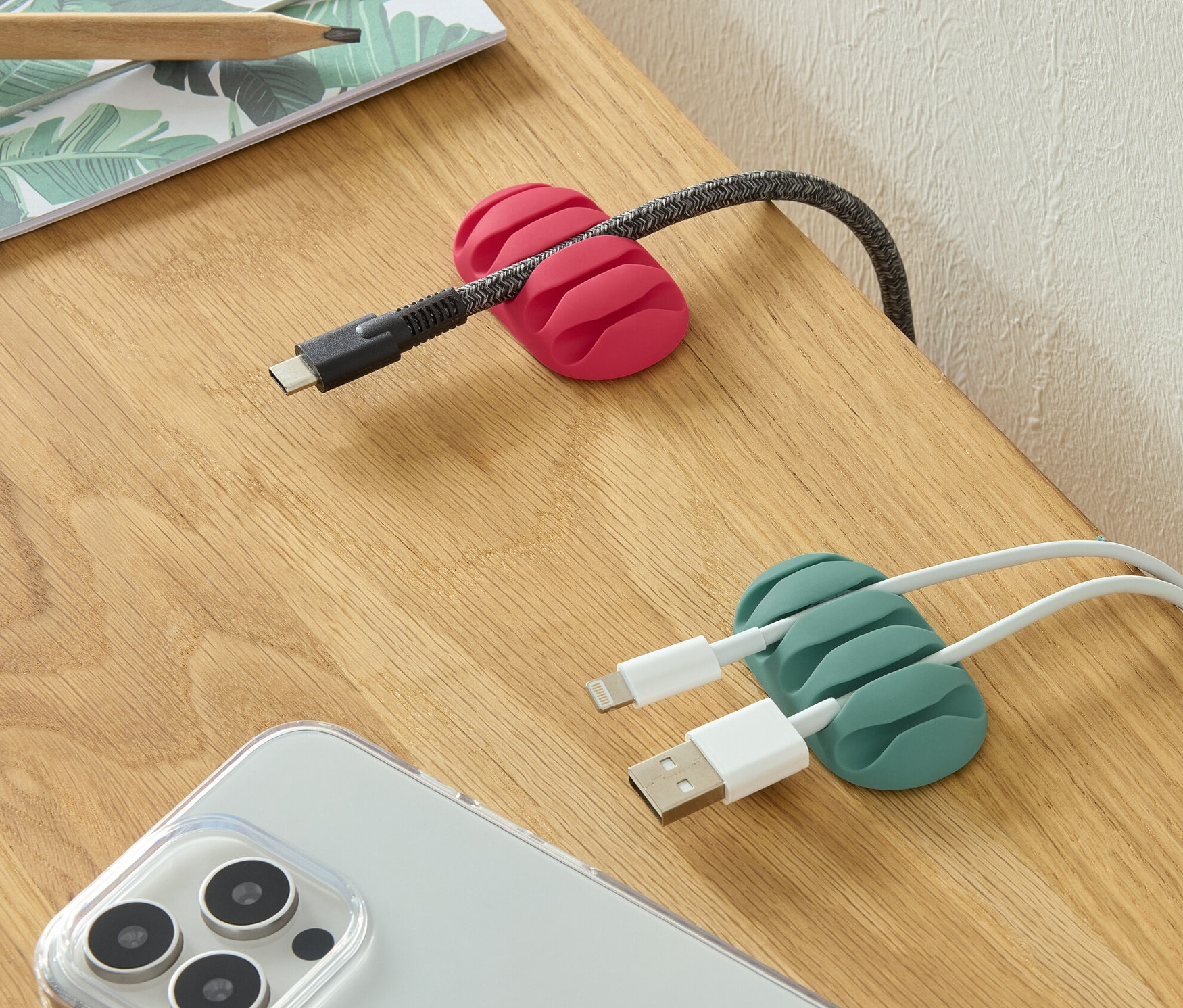 Na stole leží dva univerzální organizéry kabelů, jeden růžový a jeden zelený, spolu s telefonem a tužkami.