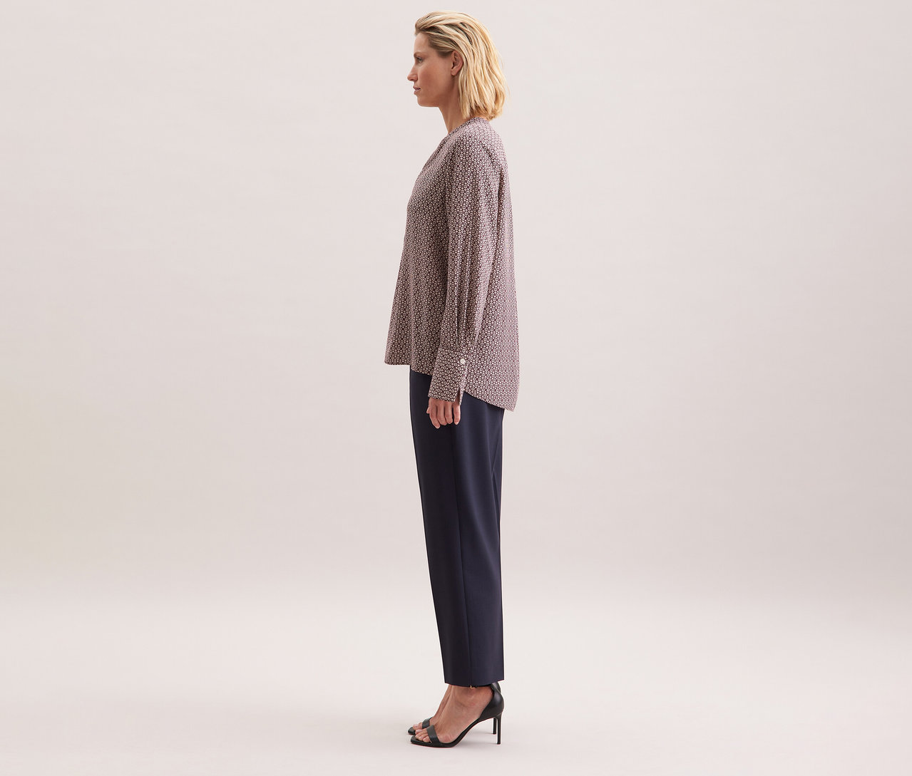 Žena má na sobě tuniku z viskózy Seidensticker s celoplošným potiskem v ecru a burgundské barvě, v kombinaci s tmavými kalhotami a černými botami na vysokém podpatku.