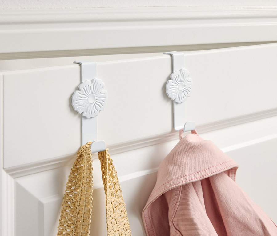Dva bílé háčky s květinovým designem zavěšené na bílých dveřích, na kterých visí žlutá taška a růžová bunda.