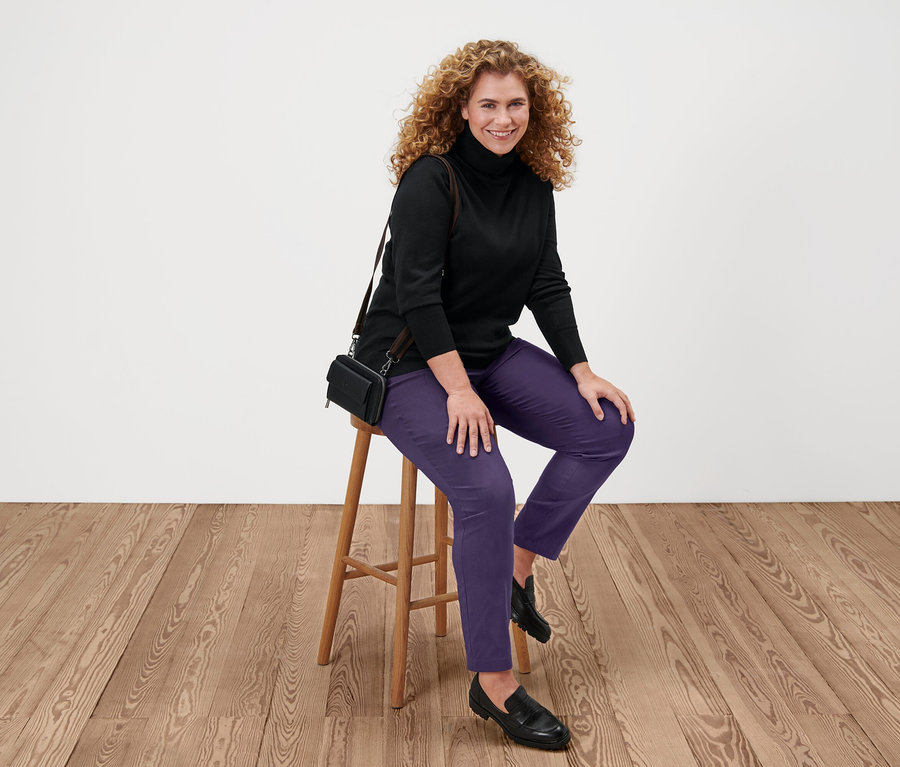 Žena sedí na stoličce a má na sobě černý rolák z jemného úpletu, fialové kalhoty a černé boty.