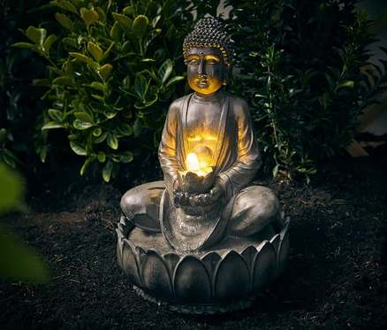 Zahradní fontána Buddha sedí na zemi před keřem.