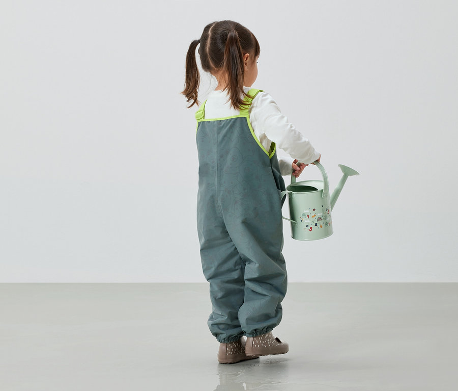 Holčička v šedých kalhotách drží v ruce zelenou konev.