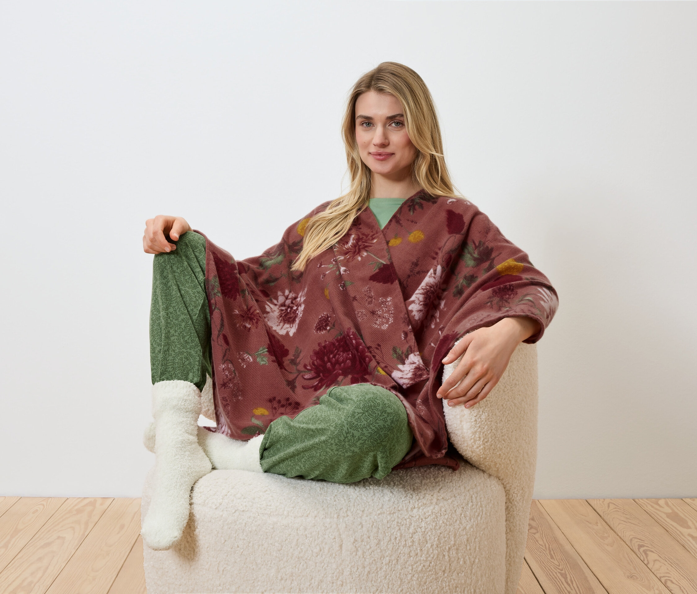 Blonďatá žena sedí v křesle, má na sobě květinové kimono, zelené kalhoty a bílé ponožky.