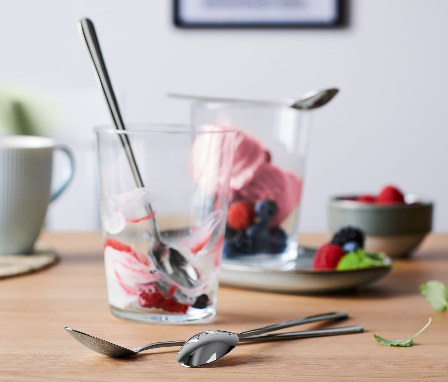 Na stole jsou dva sklenice s zbytky dezertu a tři dlouhé lžičky, 4 ks. V pozadí je hrnek a miska s ovocem.