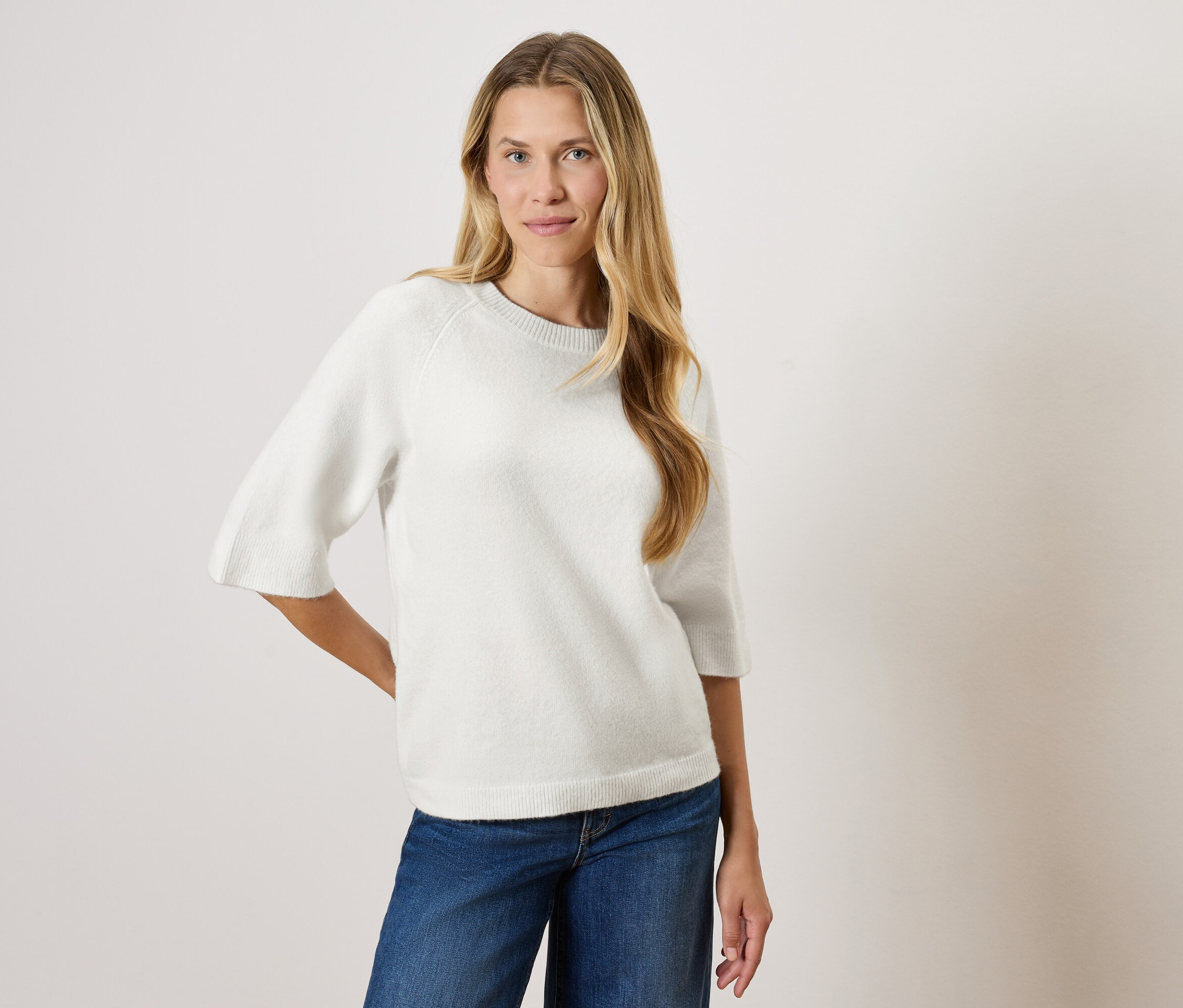 Žena s dlouhými blond vlasy pózuje v bílém svetru s tříčtvrtečními rukávy a modrých džínách.