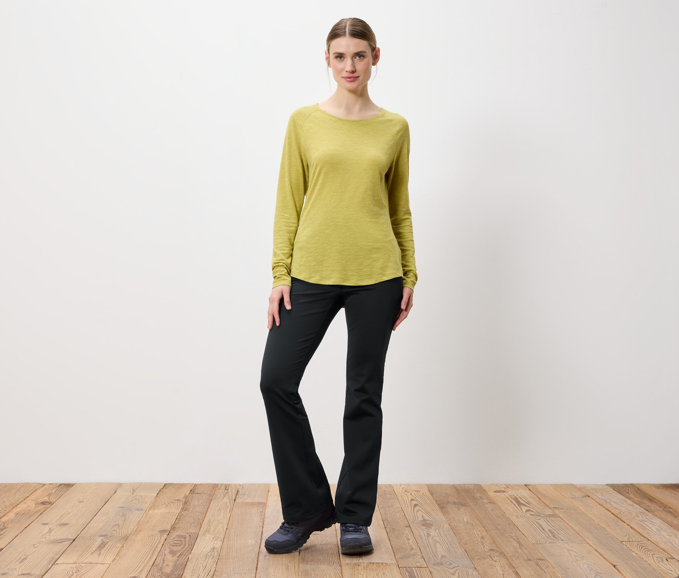 Žena pózuje ve světle zeleném tričku s dlouhým rukávem a černých kalhotách.
