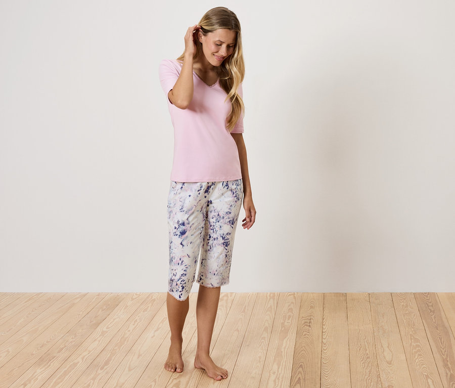 Žena v růžovém tričku a květinových kalhotách stojí na dřevěné podlaze.