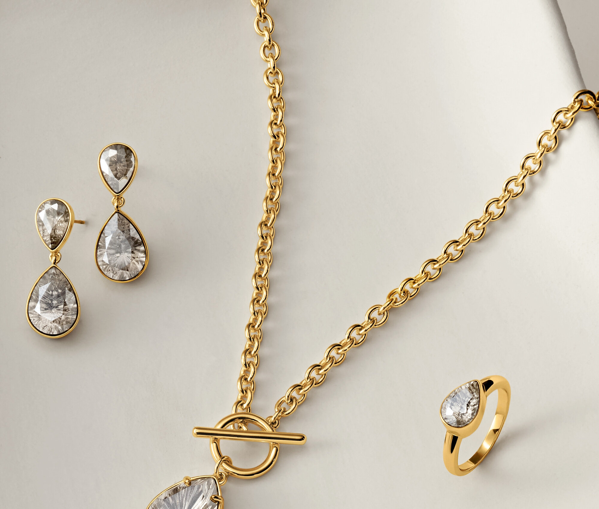 Zlatý náhrdelník, náušnice a prsten s čirými kameny ve tvaru slzy na bílém pozadí.