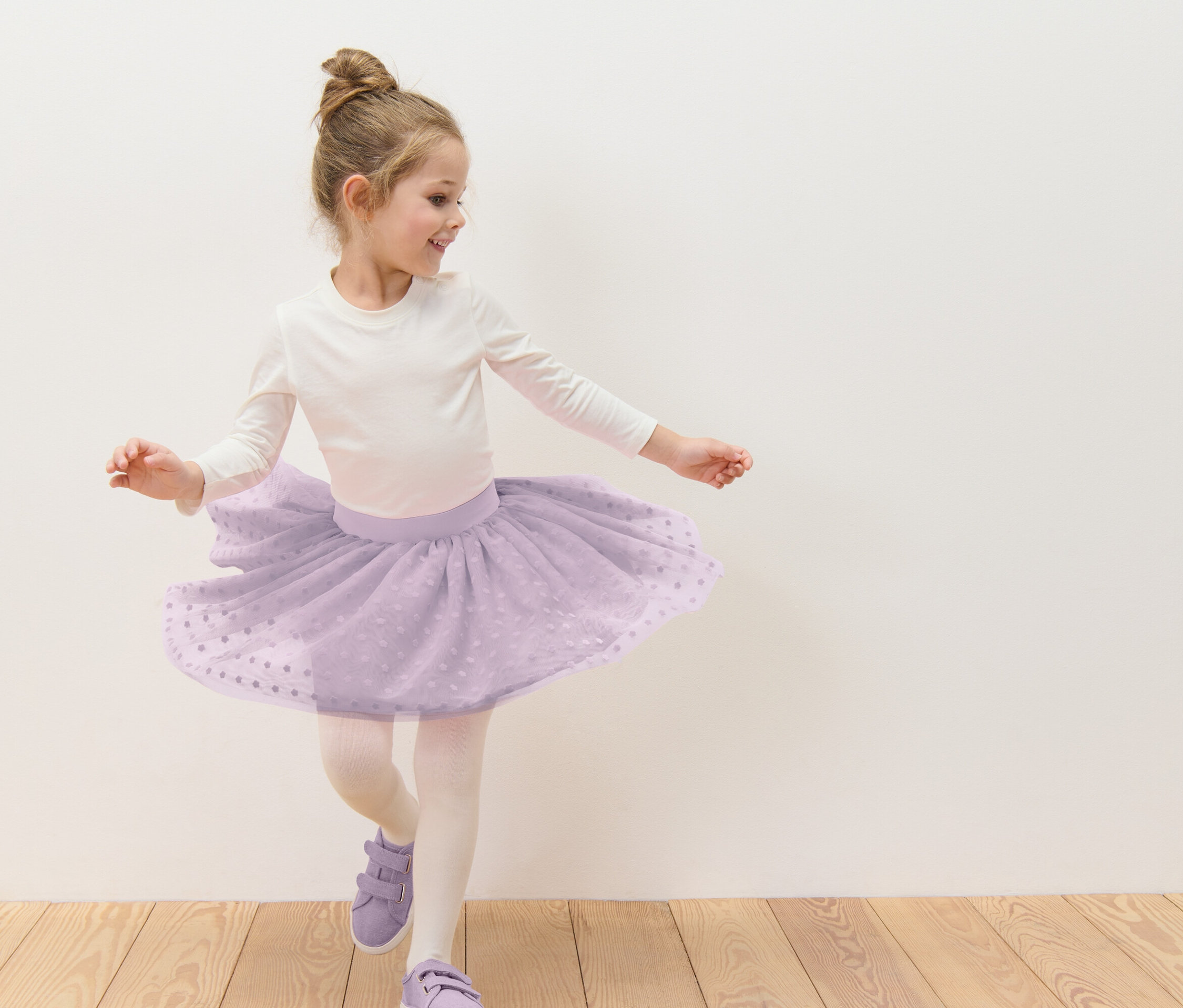 Dívka s drdolem tančí v lila tylové sukni s bílými puntíky, bílém tričku s dlouhým rukávem, punčocháčích a lila botách před bílou zdí.