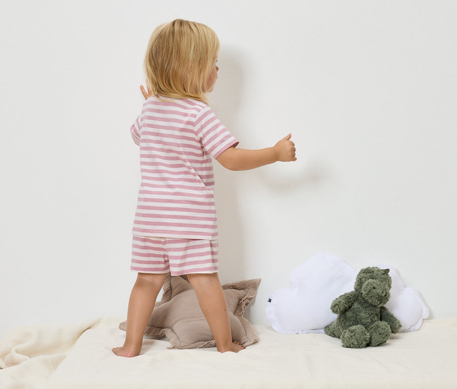 Dítě s blond vlasy v růžovo-bílém pruhovaném pyžamu stojí na béžové dece vedle zeleného plyšového dinosaura a bílého obláčkového polštáře.