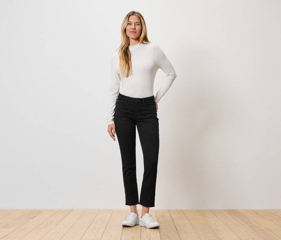 Žena stojí před stěnou, má na sobě tričko s dlouhým rukávem, černé džíny a bílé tenisky.