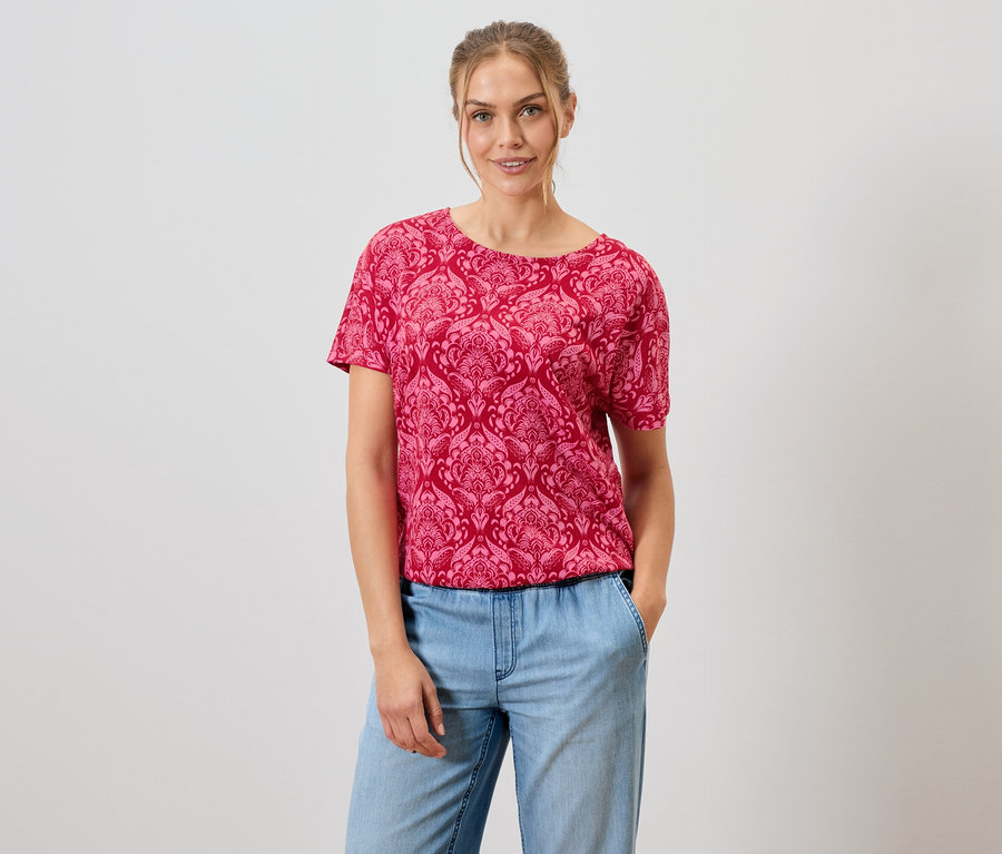 Žena má na sobě červené vzorované tričko a modré džíny.