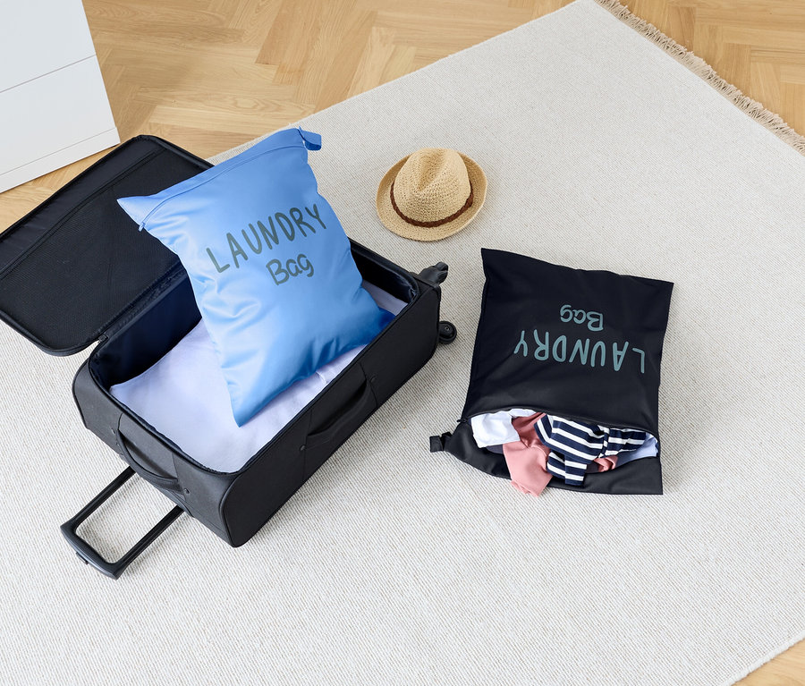 Na koberci stojí otevřený kufr s modrým vakem na prádlo uvnitř, vedle černého vaku na prádlo a klobouku.