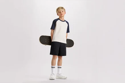 Blonďatý chlapec drží skateboard za zády, má na sobě tričko s tmavě modrými rukávy, šortky a tenisky.