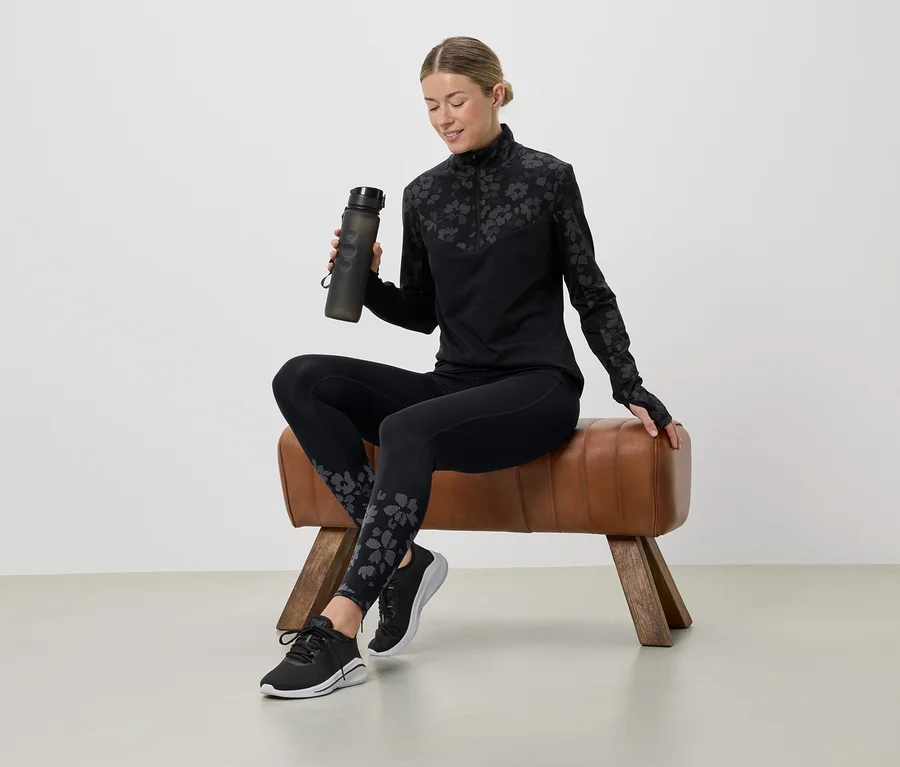 Žena sedí na koze, drží láhev s vodou a má na sobě černý sportovní outfit s květinovým vzorem.