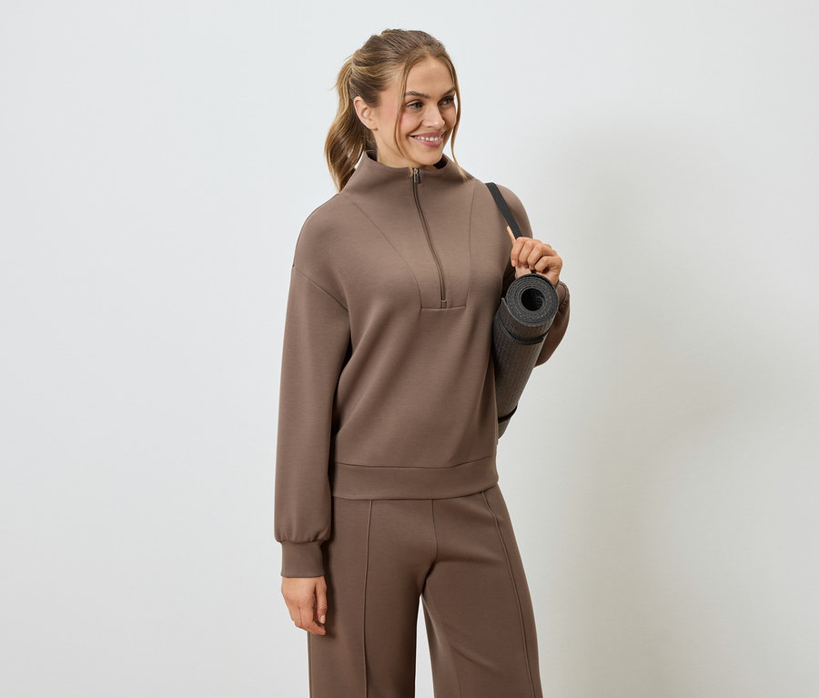 Žena v hnědé teplákové soupravě s jógovou podložkou přes rameno.