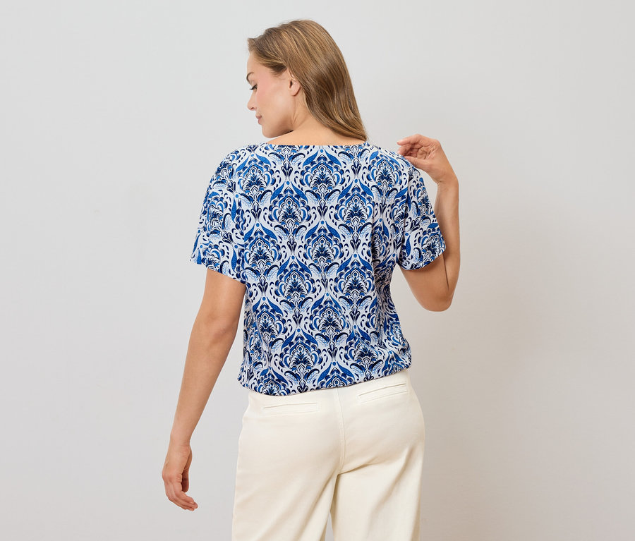 Pohled zezadu na ženu s blond vlasy v modro-bílé vzorované halence a světlých kalhotách.