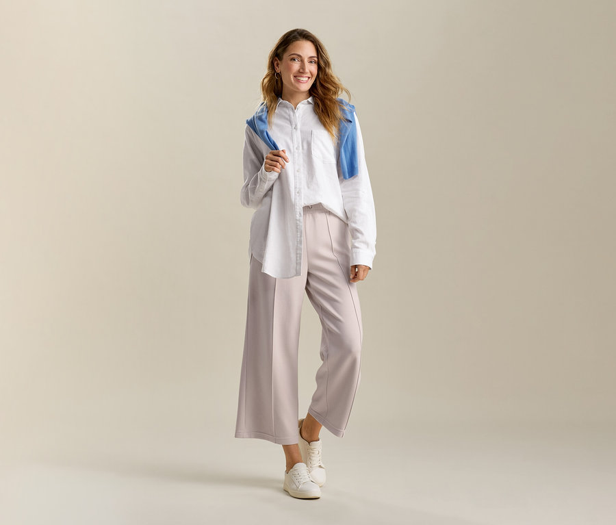 Usmívající se žena má na sobě bílou, dlouhou lněnou blůzu a pískové kalhoty culotte ze směsi modalu. Na ramenou má modrý svetr.