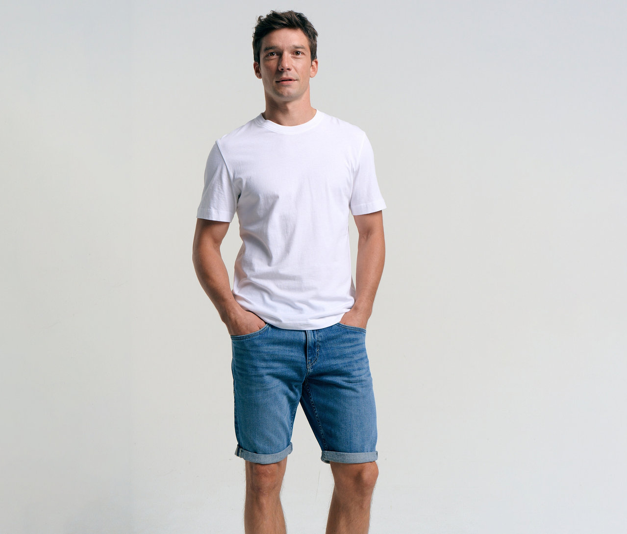 Muž v bílém tričku a džínových šortkách Tom Tailor TTJOSH REGULAR SLIM, mid stone bright blue denim, s rukama v kapsách.