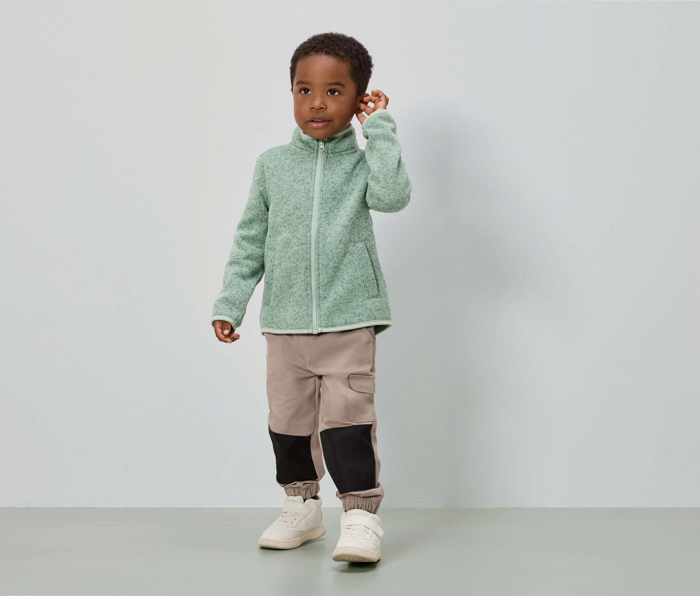 Chlapec stojí před bílou stěnou a má na sobě světle zelenou fleecovou bundu, béžové kalhoty s černými vložkami a bílé boty.