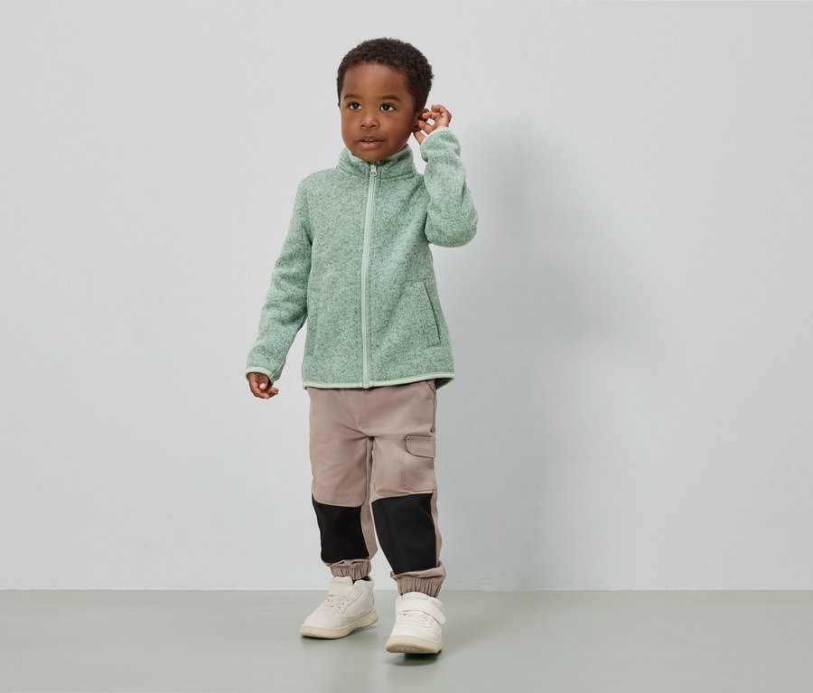 Chlapec stojí před bílou stěnou a má na sobě světle zelenou fleecovou bundu, béžové kalhoty s černými vložkami a bílé boty.