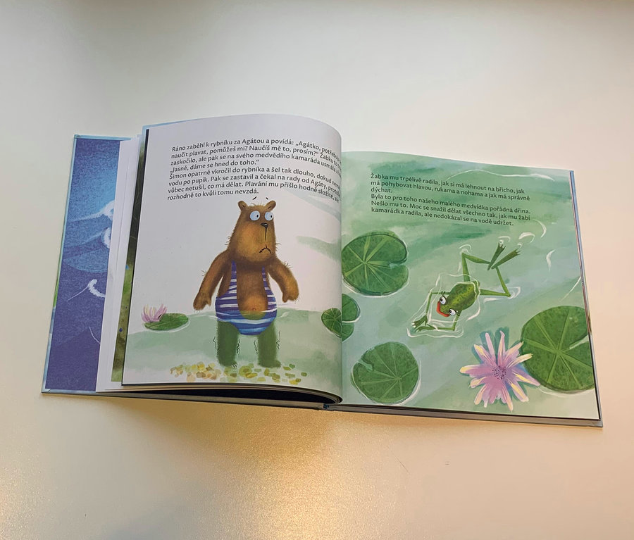 Otevřená kniha zobrazuje ilustraci medvěda v plavkách a žáby v rybníku.