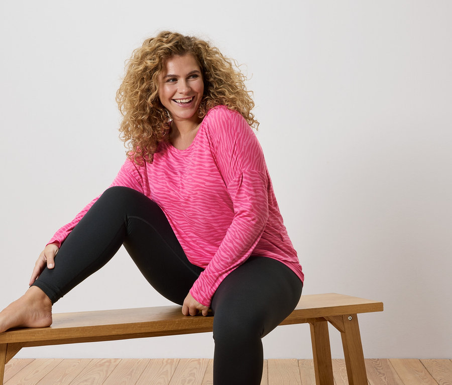 Žena s kudrnatými vlasy sedí bosá na dřevěné lavičce, má na sobě růžové tričko s dlouhým rukávem se vzorem zebry a černé legíny.