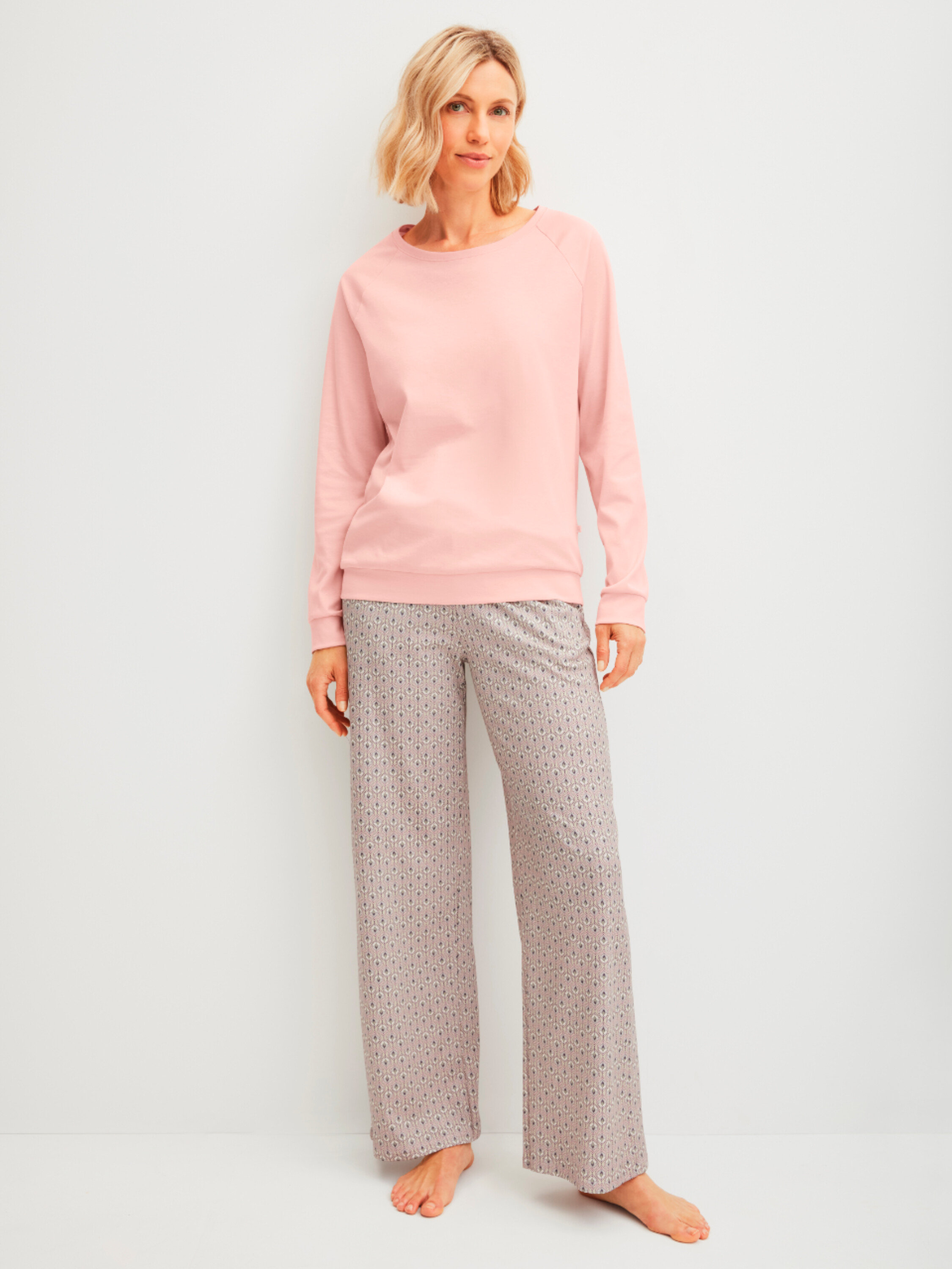 Žena stojí bosá v růžovém svetru a vzorovaných kalhotách před bílou stěnou.