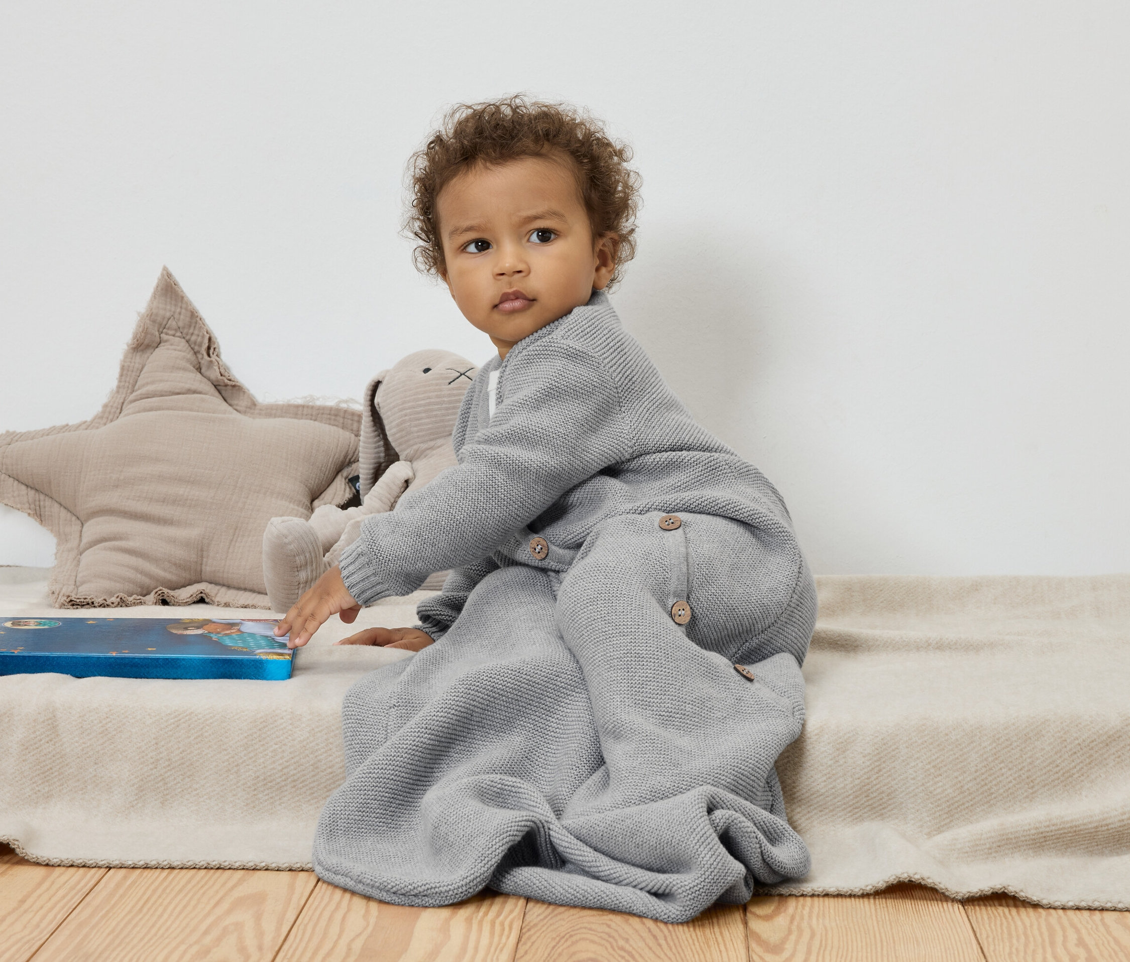 Dítě sedí na béžové dece vedle plyšové hvězdy a králíka a dotýká se modré knihy.