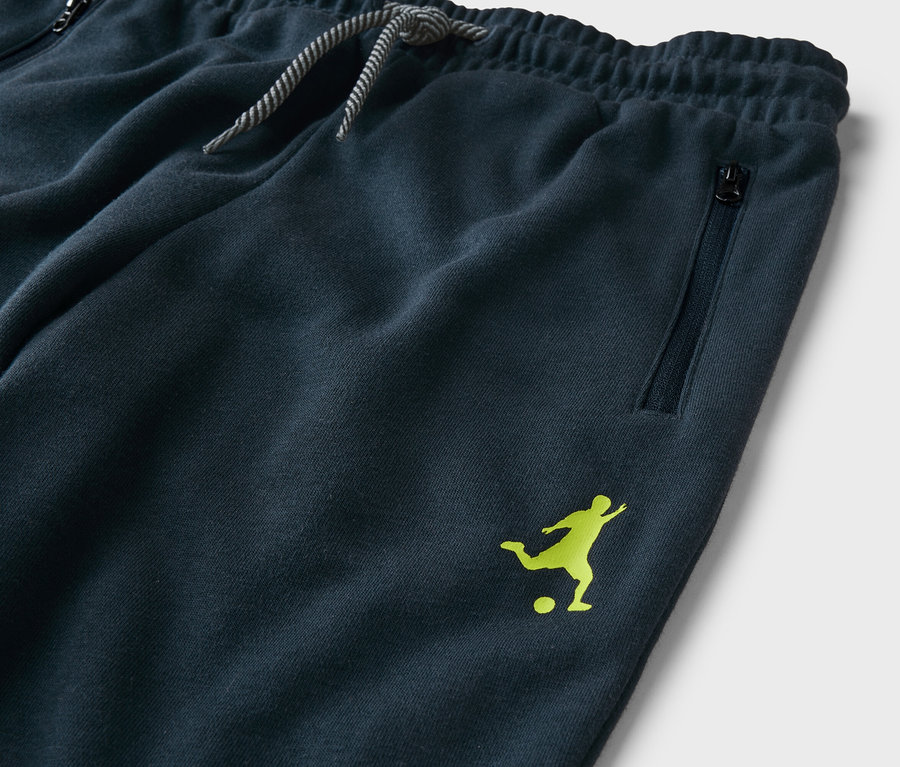 Detailní záběr na tmavě modré tepláky se stahovací šňůrkou, kapsou na zip a světle zeleným logem fotbalisty.