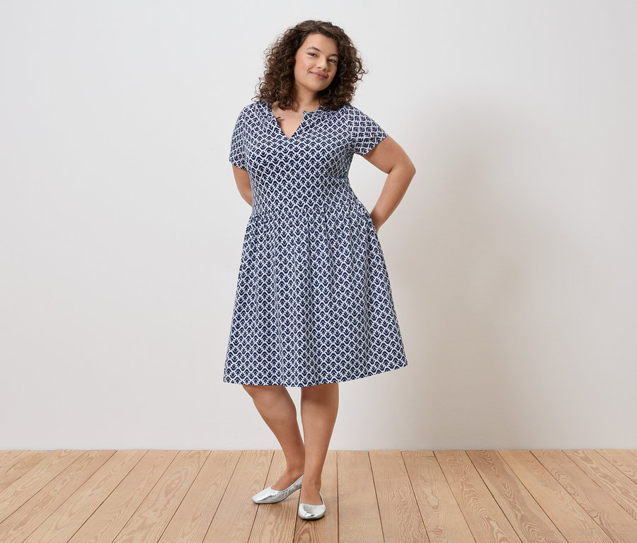 Žena s kudrnatými vlasy v modro-bílých šatech a stříbrných botách stojí s rukama v kapsách před zdí.