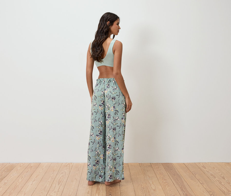 Žena stojí zády ke kameře a má na sobě světle zelený top a volné květinové kalhoty.