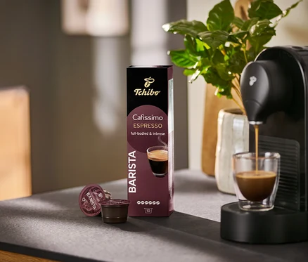 Espresso tekoucí do sklenice z kávovaru Cafissimo BARISTA vedle balení 10 kapslí.