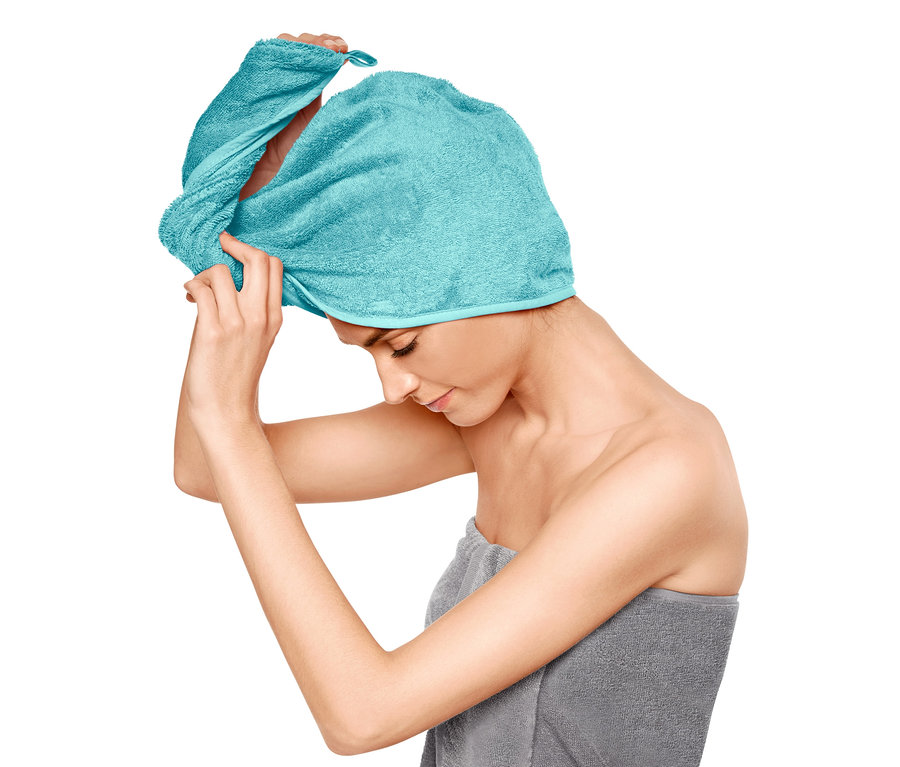 Žena si upravuje ručníkový turban na hlavě, tělo má ovinuté šedým ručníkem.