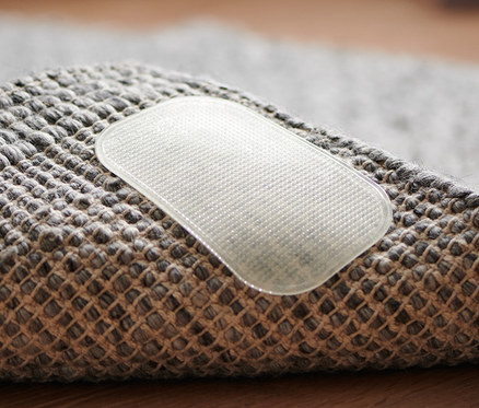 Detailní záběr na rohovou část šedého koberce s jednou průhlednou protiskluzovou podložkou, 4 ks.