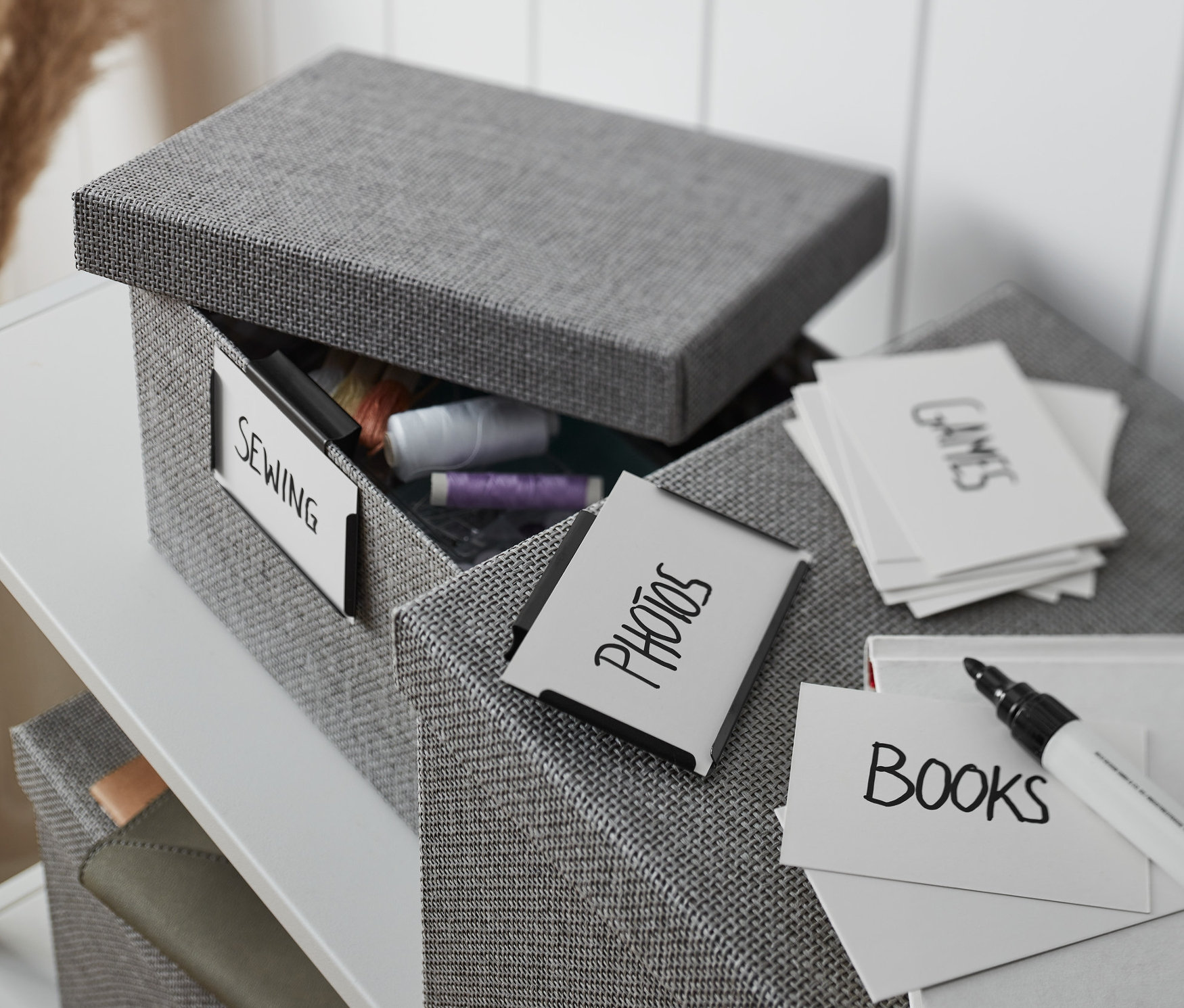 Otevřený šedý látkový box s kovovými svorkami, štítkem "šití" a obsahem nití, vedle boxu s kartami s nápisy.