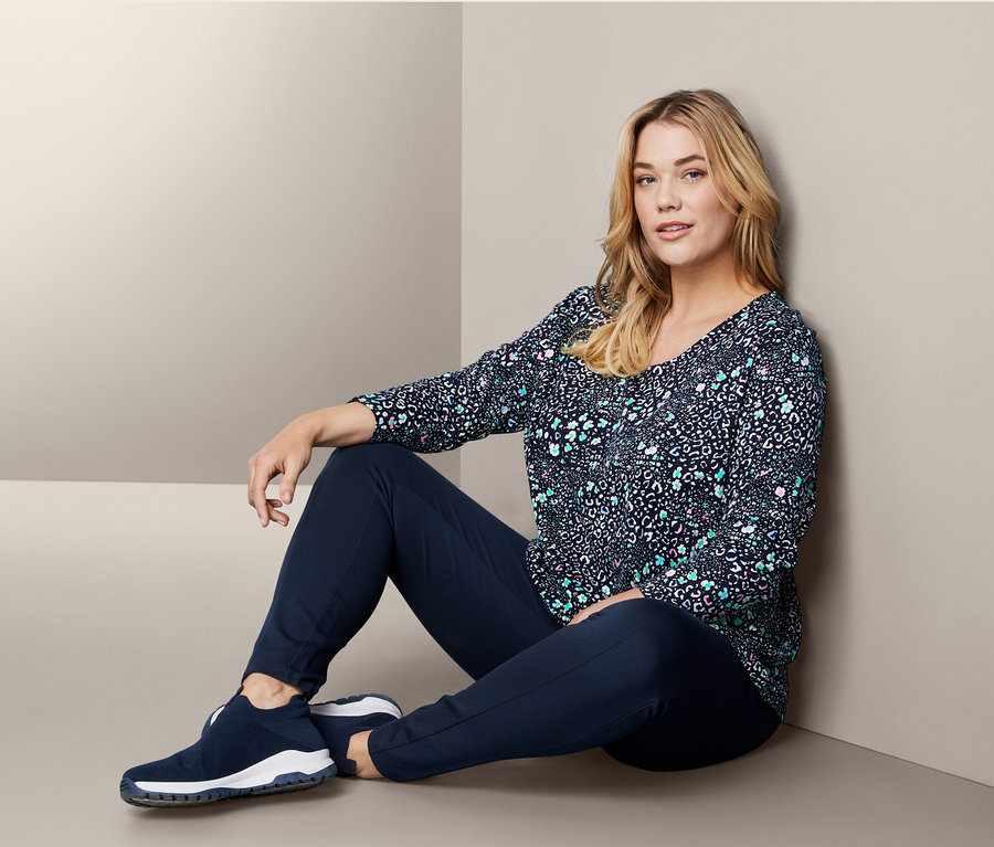 Žena sedí na zemi a má na sobě boty sneaker, triko s dlouhým rukávem a tvarující žerzejové kalhoty.