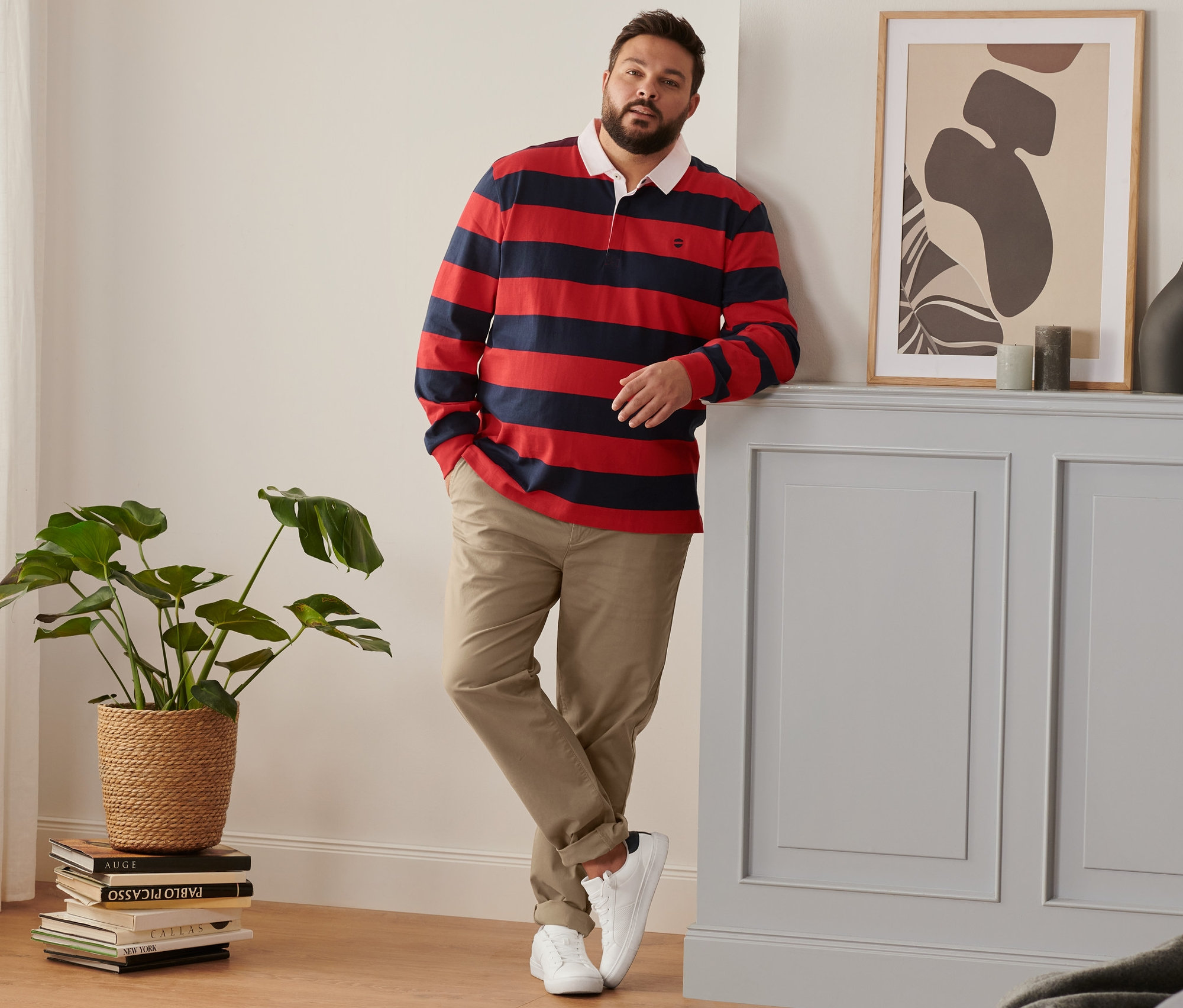 Muž opřený o zeď má na sobě boty sneaker, kalhoty chino barvy velbloudí srsti a tmavomodro-červenou polokošili s dlouhým rukávem.