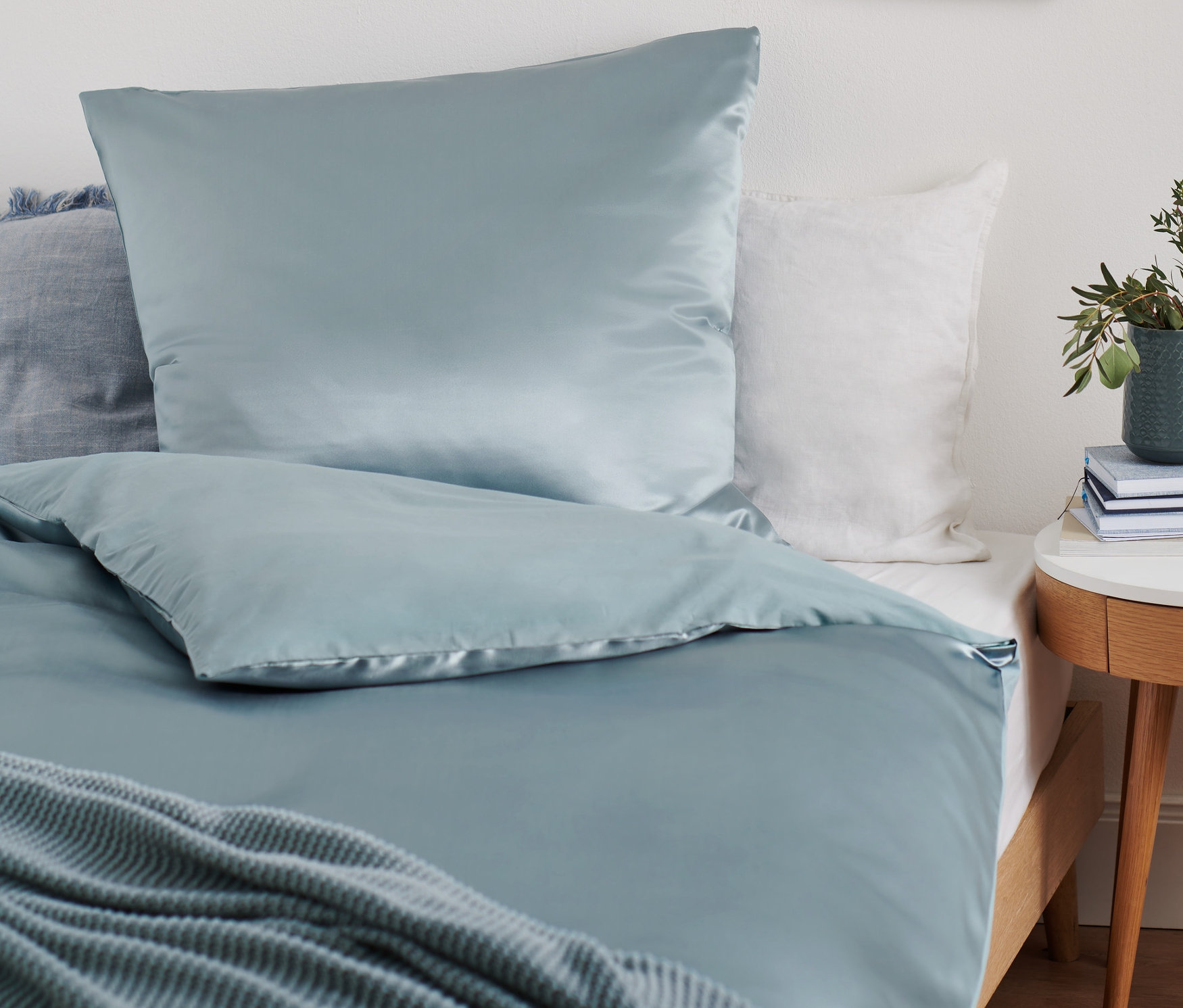 Světle modré prémiové povlečení z hedvábí a bavlny standardní velikosti na posteli. Vedle postele stojí stolek s rostlinou v květináči.