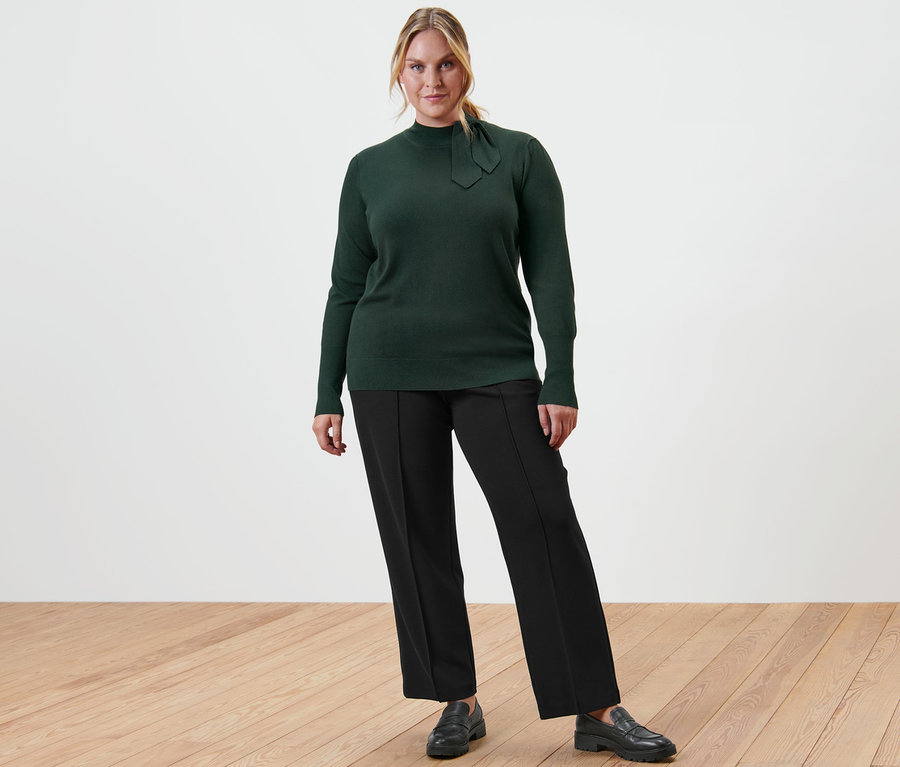 Žena pózuje v zeleném svetru z jemné pleteniny s mašličkovým detailem, černých žerzejových kalhotách s puky a kožených mokasínech.