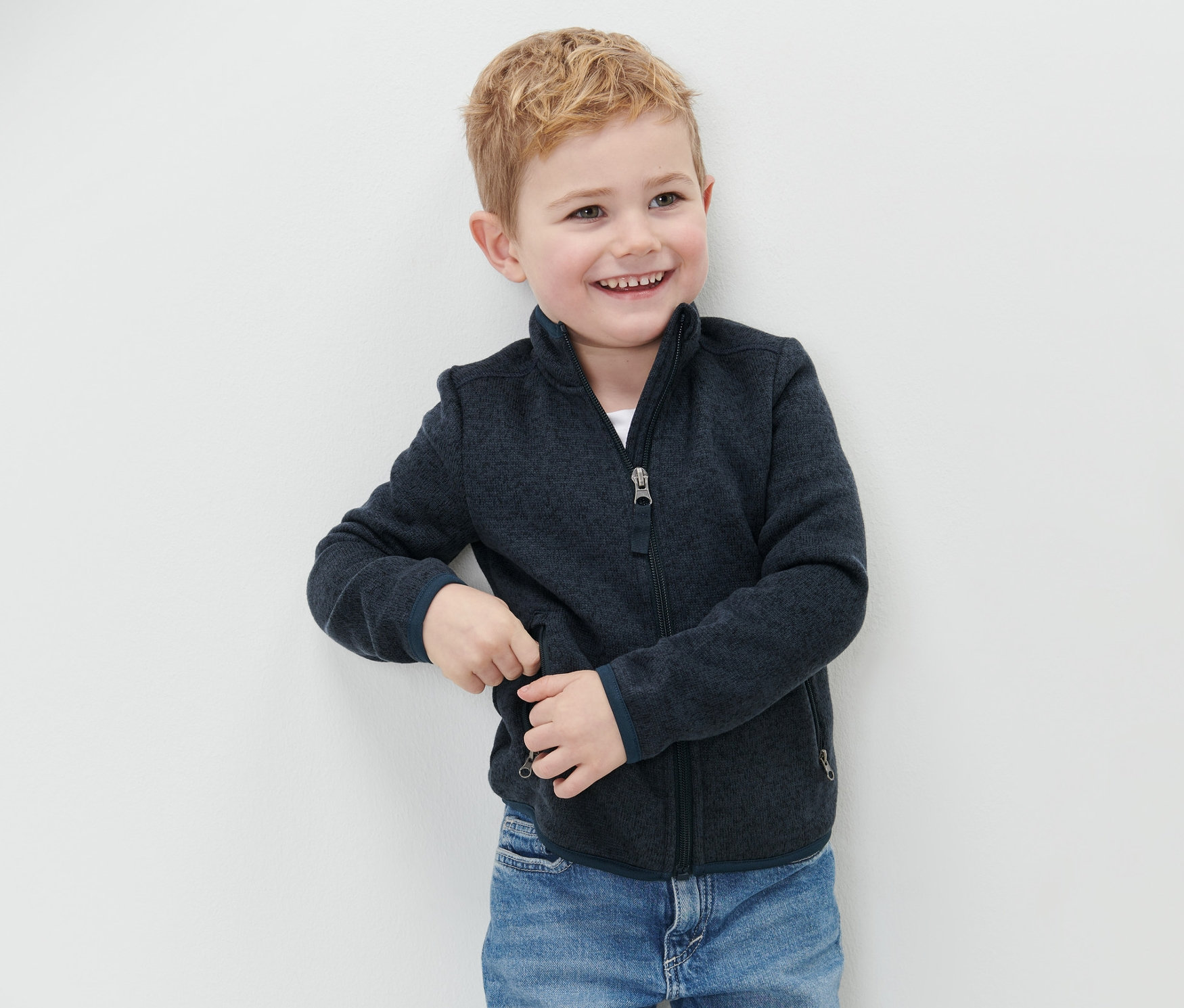 Chlapec se usmívá a stojí u zdi v dětské mikině z pleteného fleecu a modrých džínách.