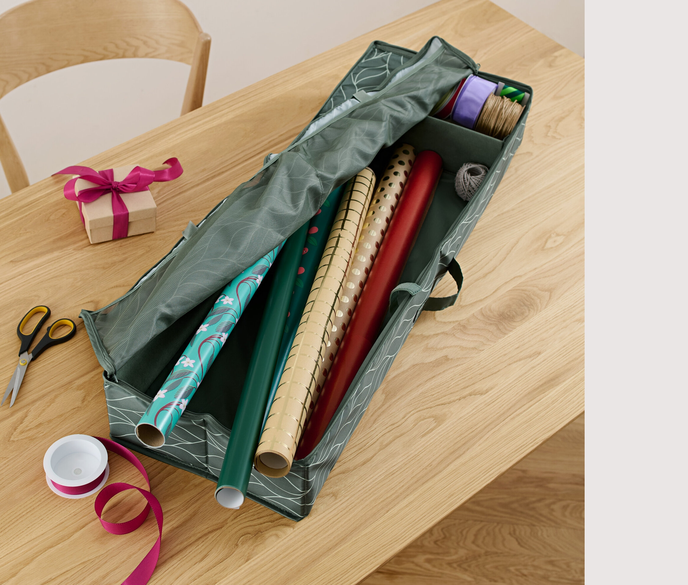 Na stole leží organizér na balicí papír, nůžky, stuha a zabalený dárek.