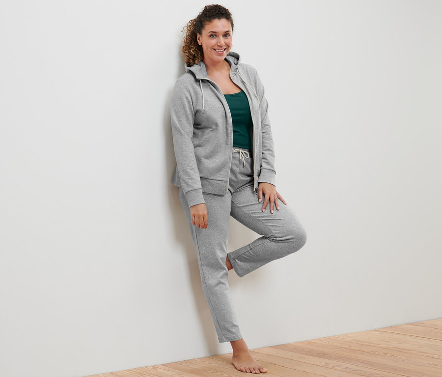 Žena opřená o stěnu má na sobě šedou mikinu, zelený top a šedé teplákové kalhoty.
