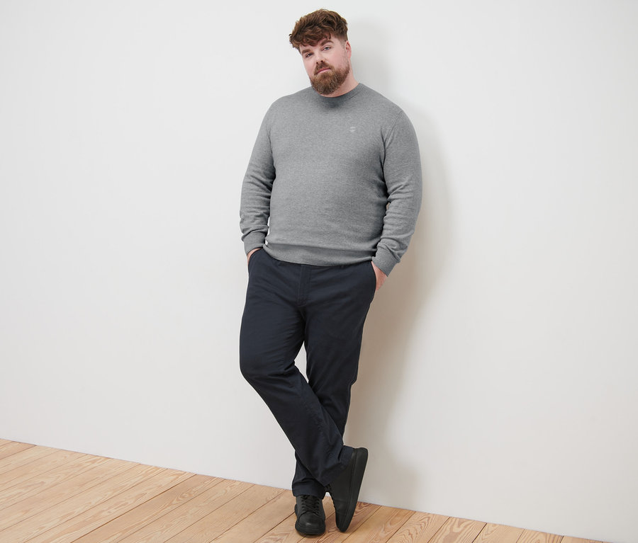 Muž se opírá o stěnu a má na sobě šedý svetr s kulatým výstřihem a námořnické modré kalhoty chino.
