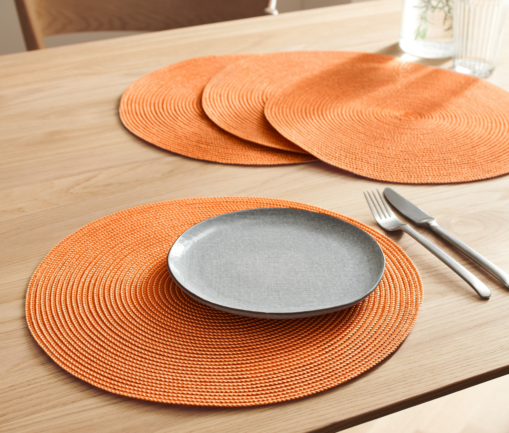 Na stole leží 4 oranžové prostírání. Na jednom z nich je šedý talíř, nůž a vidlička.