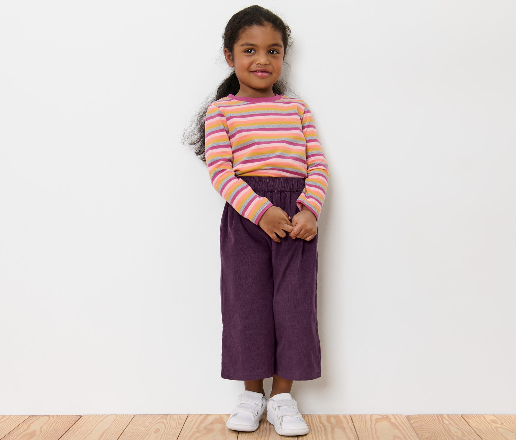Dívka stojí a má na sobě dětské kalhoty culotte, pruhované tričko a bílé boty.