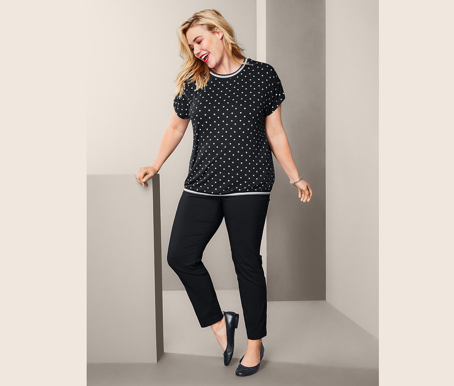 Žena pózuje v tričku s krátkým rukávem a kopretinovým vzorem, strečových kalhotách s délkou po kotníky a kožených balerínách.