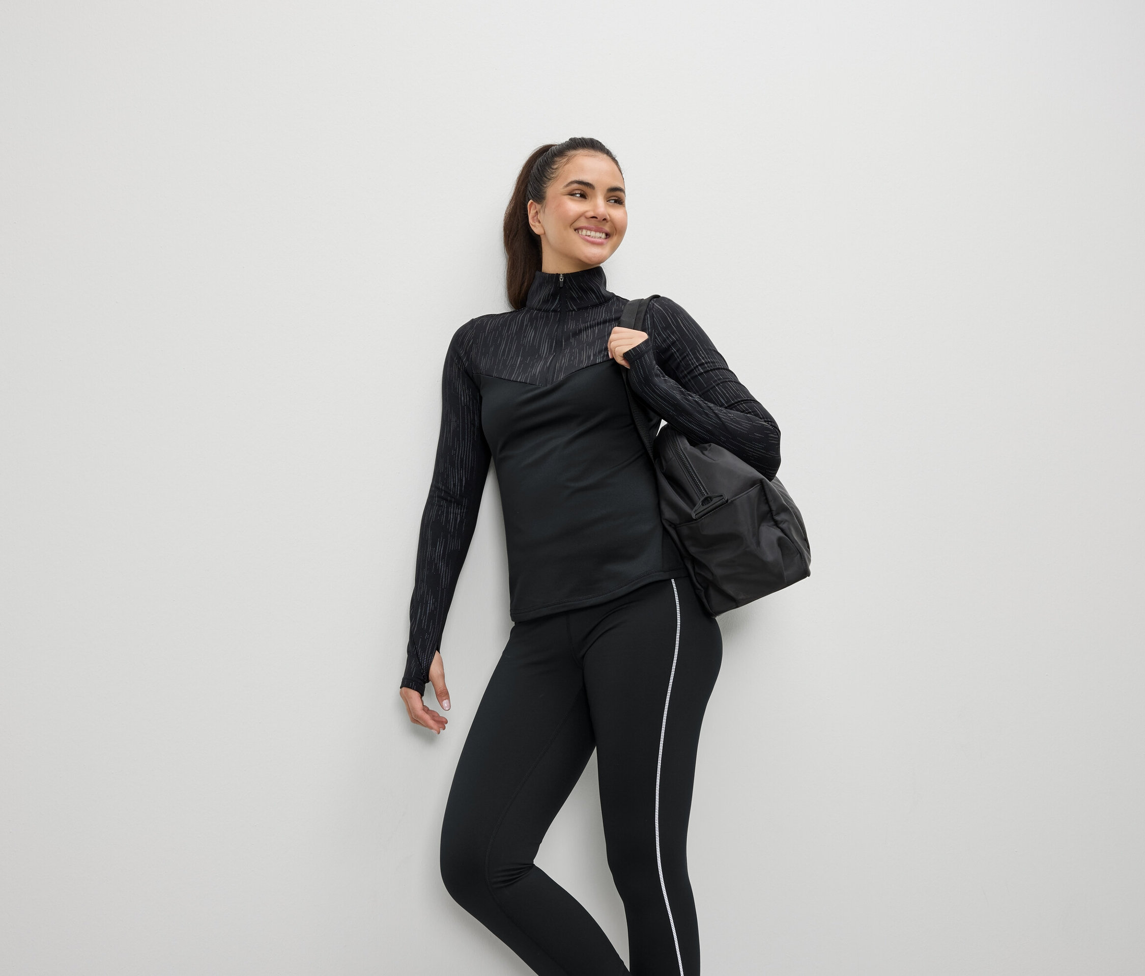 Usmívající se žena v černém sportovním oblečení s taškou přes rameno.