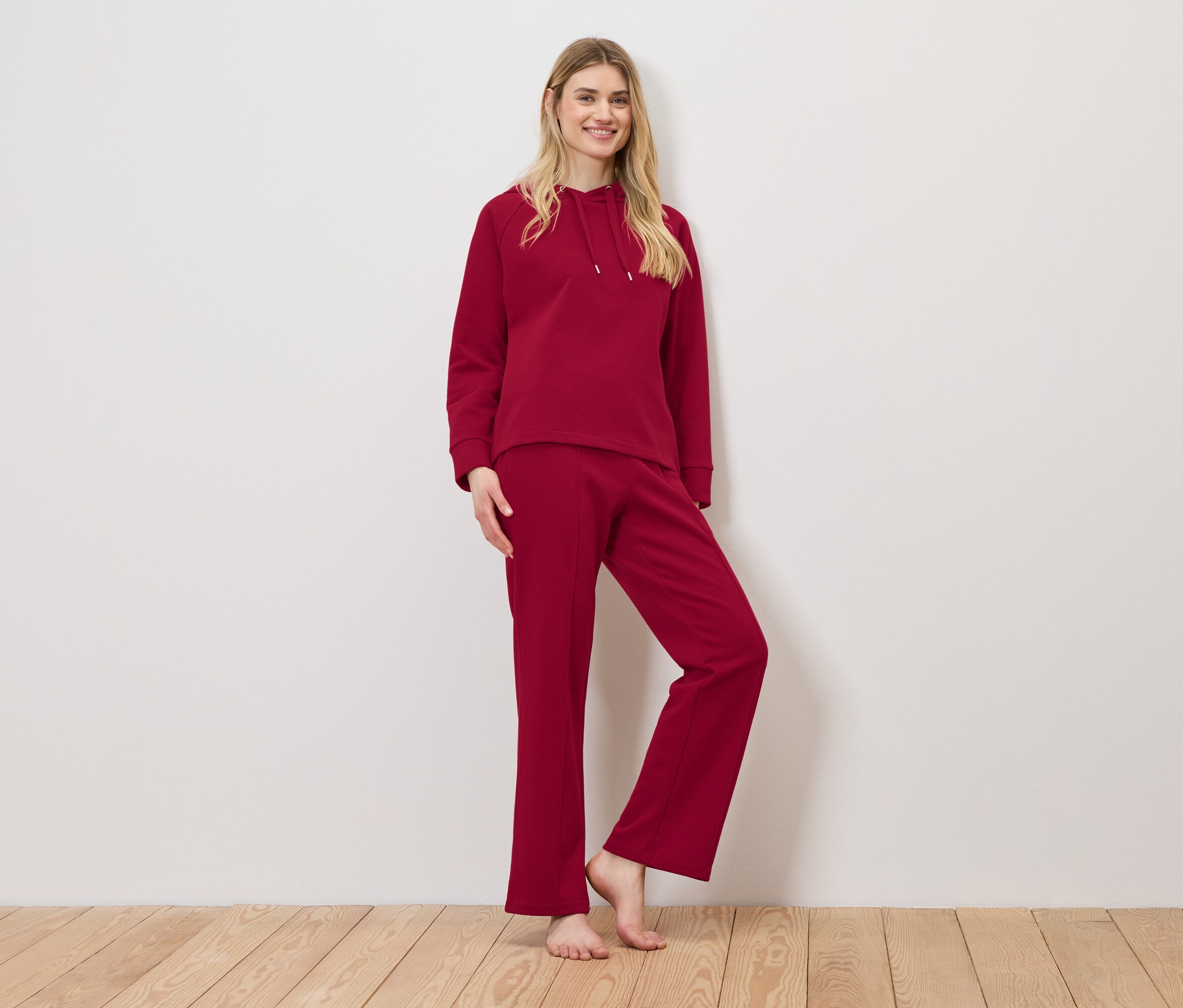 Žena pózuje v červené teplákové soupravě skládající se z mikiny s kapucí a tepláků.