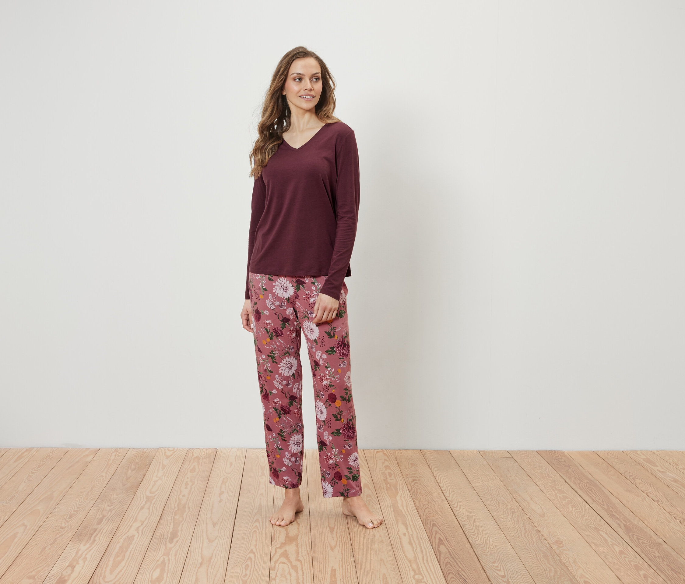 Žena stojí na dřevěné podlaze a má na sobě bordó tričko s dlouhým rukávem a růžové květinové kalhoty.